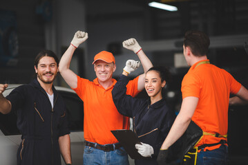Group of young and senior male and female car mechanics wearing uniform with cap and gloves in garage enjoying and celebrating with dance and smiling with hands up while holding clipping board