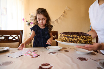 Charming Caucasian birthday girl in trendy navy dress with dots, plays with candles while her mom brings a homemade festive chocolate cake on the table. Celebrating fifth anniversary. Birthday party