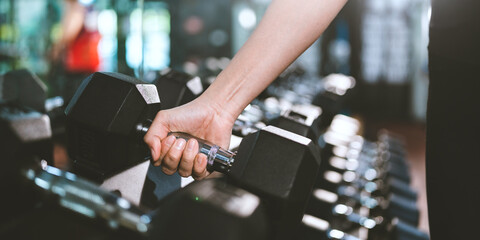 Closeup of hands of young fit and healthy female in sportswear lifting single hand weights from rack with multiple dumbbells while practicing workout for arms in modern gym and fitness club