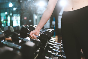 Closeup of hands of young fit and healthy female in sportswear lifting single hand weights from rack with multiple dumbbells while practicing workout for arms in modern gym and fitness club