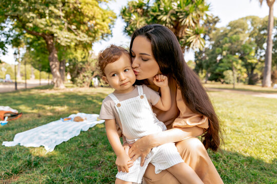 Spectacular lovey brunette woman with long dark hair is holding her adorable little daughter on knees and kissing in the park in sunny warm day. 