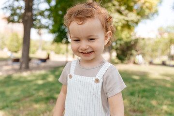 Happy excited lovely kid with curls wearing summer white suit walking in park in sunny warm day. Happy girl relaxing on spring or summer grass outdoor. Cute child having fun outdoor