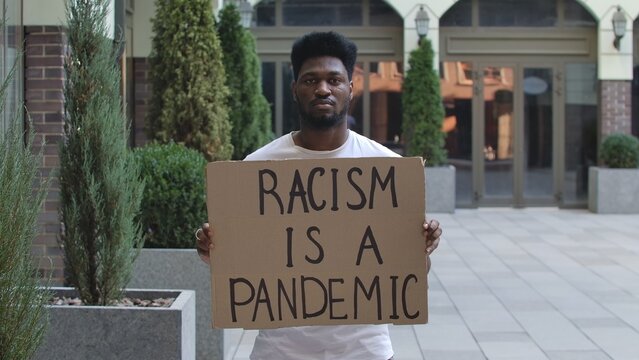 Young African American Man Stands With A Cardboard Poster RACISM IS A PANDEMIC In Public Outdoor Place. An Anti Racist Movement To Protest Against Injustice And Police Brutality. Street Demonstration.