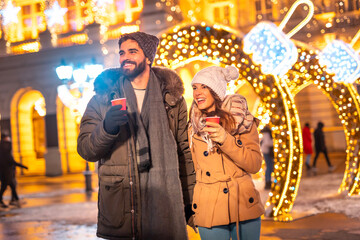 Couple drinking mulled wine and walking down decorated city streets while celebrating Christmas