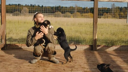 Concept adoption of abandoned pets. Young handsome Caucasian man with dreadlocks came to dog shelter to choose new friend. Volunteer plays with puppies in Alaskan husky kennel in fall. - Powered by Adobe