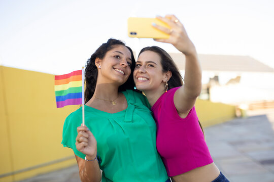 A beautiful lesbian young couple embraces and holds a rainbow flag. Girls taking selfie photo..