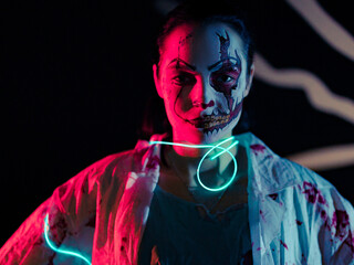 Young woman with glowing led band portrays bloodthirsty zombie with wounds on her face and horror teeth.