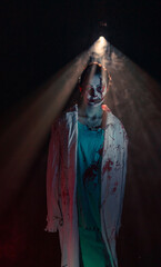 Young woman portrays bloodthirsty zombie with horror wounds and bloody clothes against dark background under light.