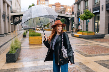 Portrait of laughing young woman in elegant hat dancing and having fun with transparent umbrella on beautiful city street, enjoying rainy weather outdoors. Concept of female lifestyle at autumn season