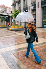 Fototapeta na wymiar Vertical back view to unrecognizable young woman in fashion hat walking on European city street with transparent umbrella enjoying rainy weather outdoors. Concept of female lifestyle at autumn season.