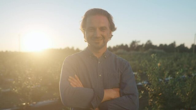 Close up of handsome happy male farmer looking at camera and smiling while stands in filed plantation. Positive man outdoors in village farmland at sunset. Farming business, Agriculture concept