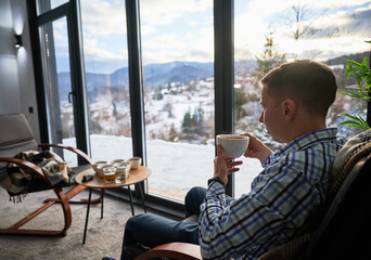 Young man enjoying weekends inside contemporary barn house. Side view of tourist sitting on chair, holding cup of tea, enjoying view of mountain landscape through panoramic windows in new cottage.
