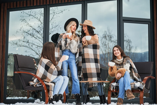 Young women enjoying winter weekends on terrace of contemporary barn house. Four girls having fun, sitting on chairs and drinking hot tea.