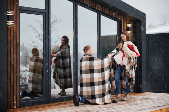 Happy woman holding bouquet of flowers and smiling while man making marriage proposal under winter snow. Couple sharing romantic moment outside scandinavian wooden house barnhouse.