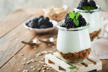 Yogurt with granola, blackberry berry fruits and muesli served in glass jar on wooden background....