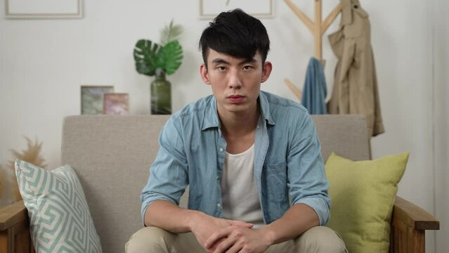 front view of a depressed asian man looking at the camera blankly on living room couch alone and feeling at a loss while staying at home during quarantine.