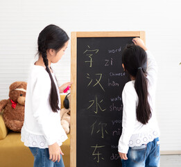 Little kid standing by blackboard happy writing Chinese text. Young Asian small girl study together at home easy learning Chinese language, write on chalkboard. Home schoolgirl lifestyle.