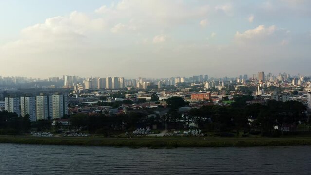 Aerial drone left trucking and panning shot of the south of S&atilde;o Paulo, Brazil from the man-made Guarapiranga Reservoir towards the Socorro neighborhood with skyscrapers and houses during sunset.