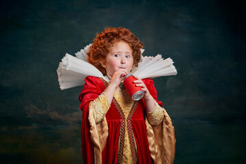 Portrait of little red-headed girl, child in costume of royal person drinking soda isolated over dark green background