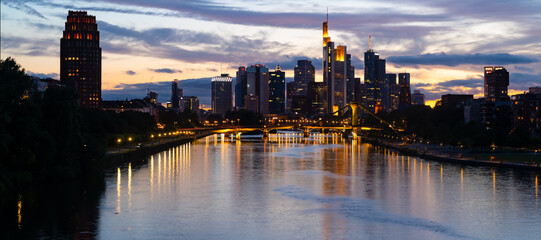 Fototapeta na wymiar Frankfurt wide angle nighttime panorama. German city with downtown skyline, tall buildings and bridge. Evening sky with clouds and reflection in River Main at blue hour with colorful illumination.
