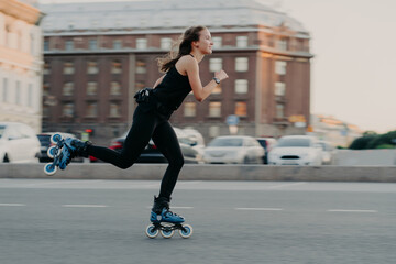 Athletic woman rides on rollers moves very fast dressed in active wear enjoys rollerblading being photographed in action poses at urban place engaged in extreme sport. Active lifestyle concept - obrazy, fototapety, plakaty