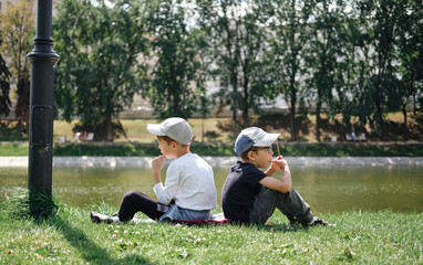 two boys sit on the grass in the park with their backs to each other and look at the lake. Summer vacation in the park