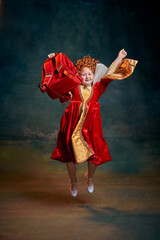 Fototapeta premium Portrait of little red-headed girl, child in costume of royal person jumping with school bag isolated over dark green background