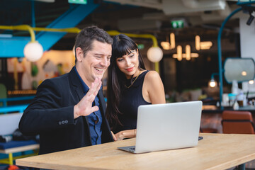 Smiling adult business man in formal clothing waving hand to someone during online video call using laptop while enjoying with wife in restaurant and office