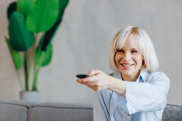 Happy young blonde woman switches channels on TV trying to choose something interesting.