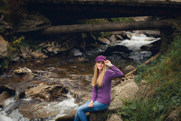 A slender young girl with long blond hair, in summer clothes and a hat, smiles and sits on the bank of a mountain river near the bridge, during the day.