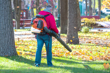 Worker operating heavy duty leaf blower in city park. Removing fallen leaves in autumn. Park...
