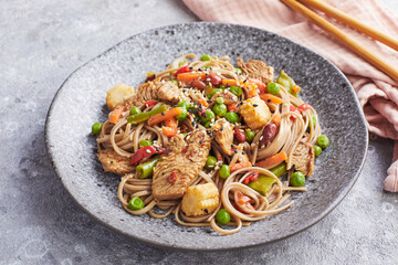 Wok with turkey meat, soba noodles, corn, green peas, green beans and carrots served on gray background with chopsticks. Asian food, concept of street food