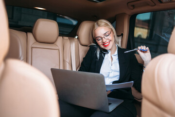 Beautiful business woman working on a laptop in a limo, smiling and talking on the phone