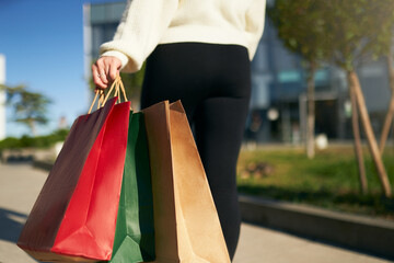 Close up of woman walking with shopping bags in hands in city from mall. Low angle view of girl going with purchases from black friday sale. Female with full paper bags. Back view.