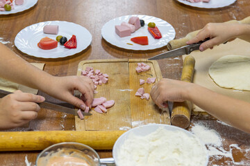 Children's hands cut pieces of sausage with knife for making pizza. Nearby are plates with vegetables and sauce.