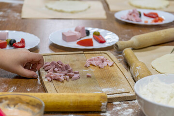 On wooden table lies cutting board with pieces of chopped sausage, plates with vegetables and kitchen utensils.