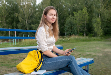 the girl sits in the stands of the school sports ground with a phone in her hands.