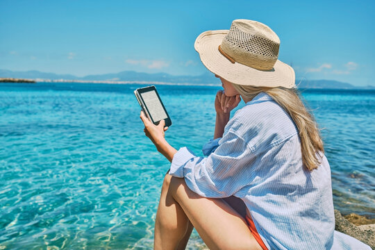 Fashion pretty woman outdoors lifestyle watching, reading on tablet ebook on the beach in summer day. Wearing wide brimmed hat, Sunbating with uv protection. Concept of beach vacation.
