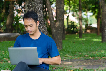 portrait of a young man remotely working outdoor casual business with laptop in the park, a student sitting on grass under tree at campus studying online technology communication education concept