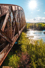 Paisaje de Puente metálico de tren o ferrocarril abandonado, oxidado y antiguo sobre un gran río con luz de medio día y el sol por encima visto desde el lado