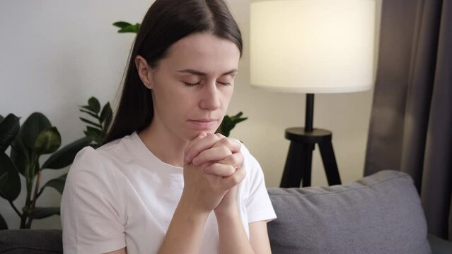 Faith in God, religion concept. Calm young woman sitting on couch with eyes closed and palms folded in prayer gesture, asking sincerely for miracle and luck pleading for innermost, express gratitude