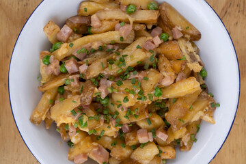 American food. Closeup view of fried potatoes with egg, ham, peas and cheese, in a white bowl on the restaurant wooden table.