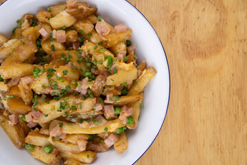 American food. Closeup view of fried potatoes with egg, ham, peas and cheese, in a white bowl on the restaurant wooden table.