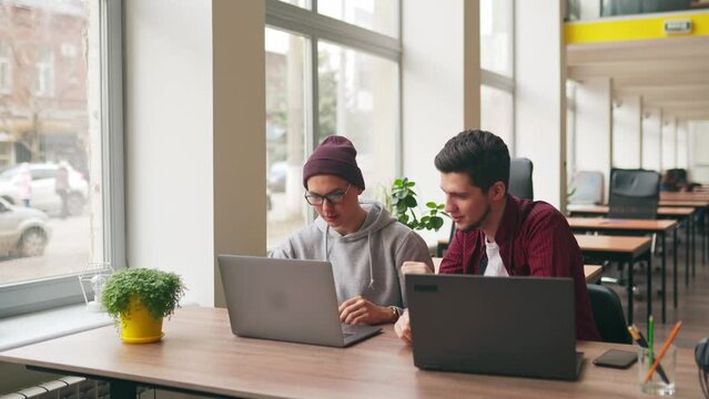 Two hipster coworkers sharing ideas about project sitting with laptops in spacious open space office. Developer discuss software program code. Designer and manager consulting on working plan.