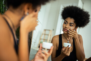 Young african american woman applying cream on face near mirror in bathroom.