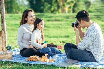Happy asian family in the garden, Father, Mother and doughter. They are having fun playing song with ukulele, reading book, using laptop in holiday.