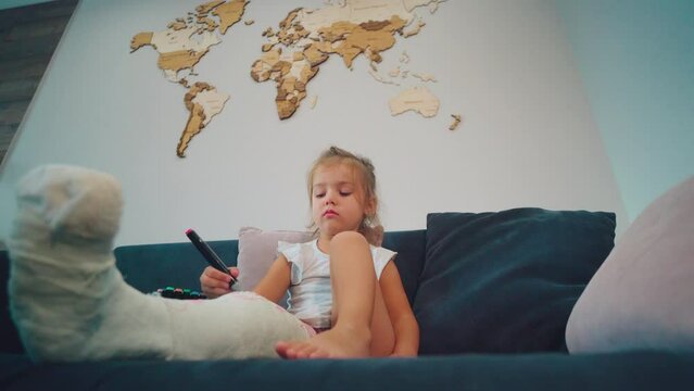 A young girl with a broken leg draws with a marker on a plaster cast. World map.