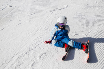 Fototapeta na wymiar tired child in a helmet, ski goggles, skis and winter overalls sits on the snow. Fell into the snow, stopped to rest. winter fun. Little boy learning to ski during winter holidays on a sunny cold day