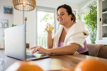 Woman drinking juice and eating at the table while watching something at the laptop