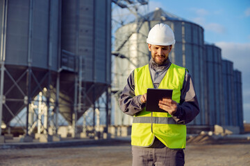 The oil and petroleum refinery worker uses tablet outside in front of the silos.